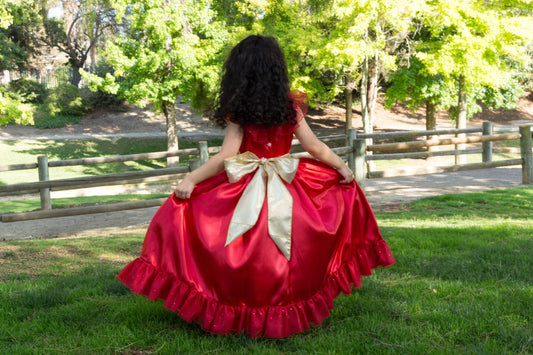 Emma's Magical Dream, Red princess ballgown with a gold tie around the waist with a big bow tied in the back and a red flower. It has a white underlay with the jewelry being gold dangle earrings. The child is twirling her dress by holding it up on both sides.